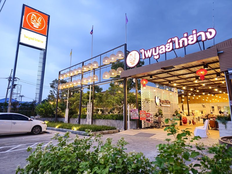 หน้าร้านไพบูลย์ไก่ย่างยามพลบค่ำ ป้ายไฟชื่อร้าน โคมไฟสานเรียงแถว และป้ายเสาสูงสีส้ม